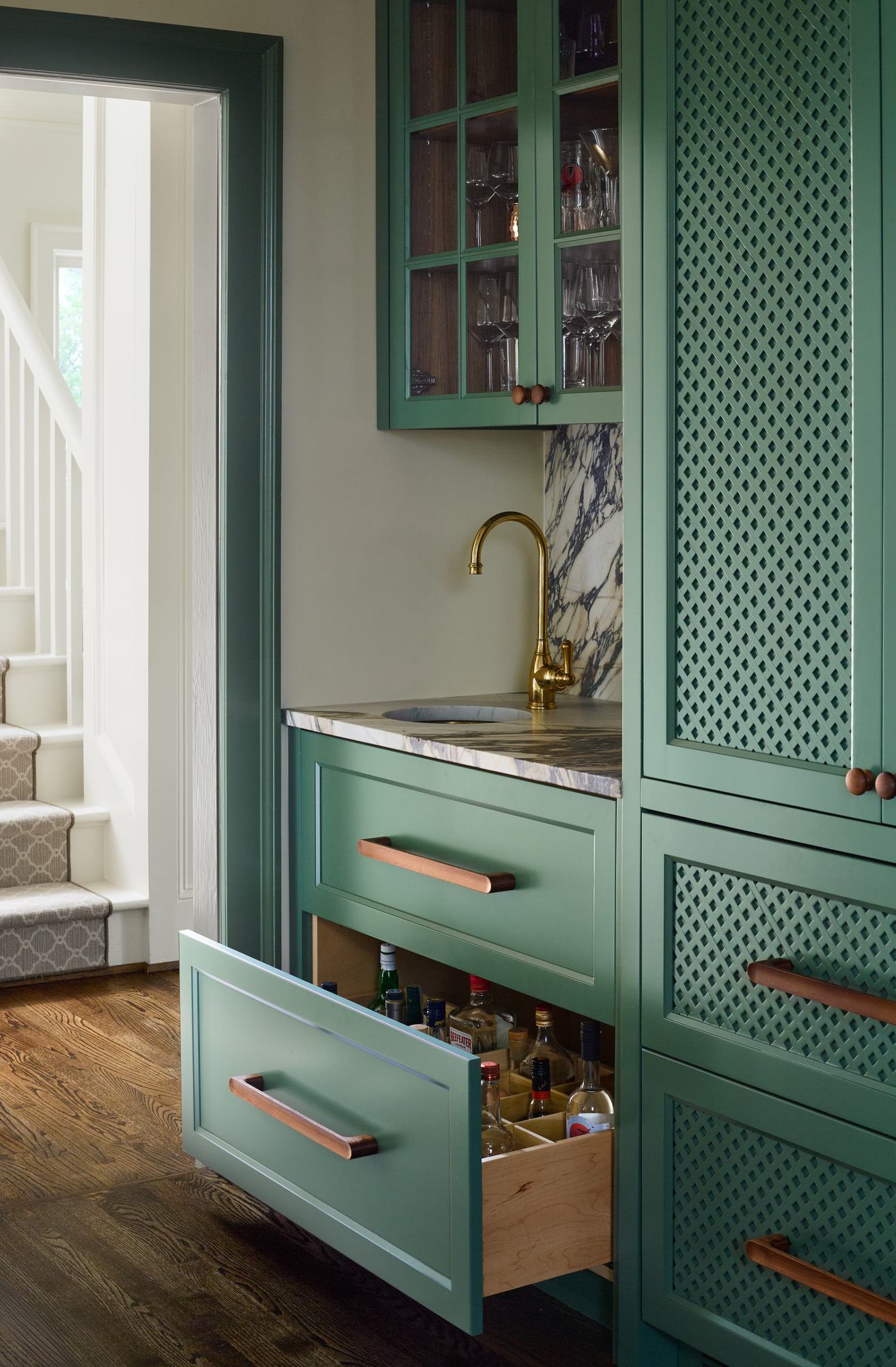 Open pantry drawer with glassware and bar bottles behind green cabinetry